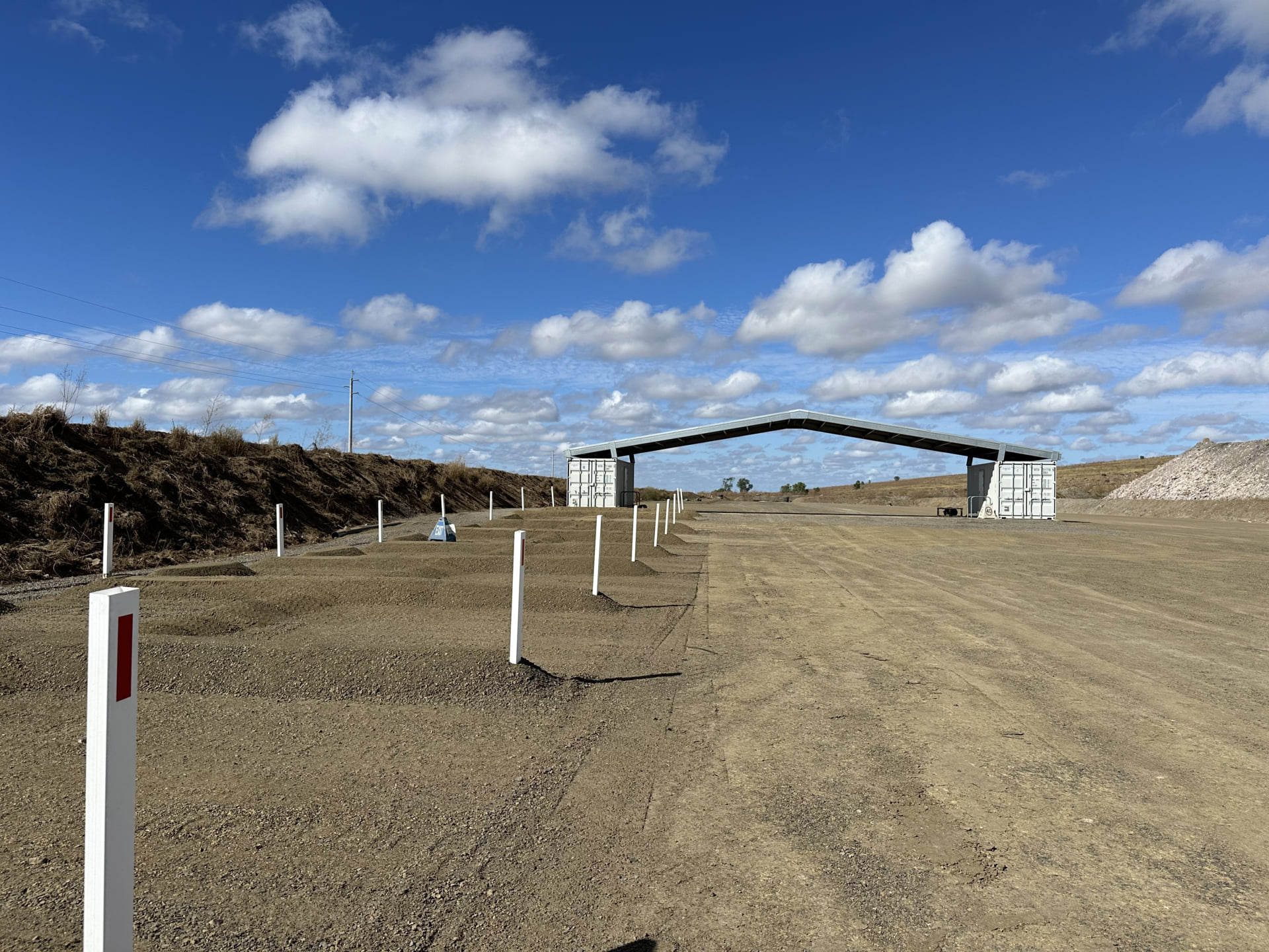A dirt roadway leading to a partially constructed shelter with a metal roof supported by two shipping containers, surrounded by marked gravel mounds and white boundary posts under a bright blue sky with scattered clouds.