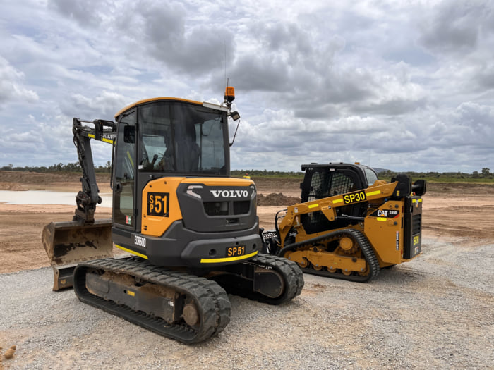 A Volvo ECR50D excavator and CAT 259D3 track loader parked on a gravel site under a cloudy sky, both labeled with fleet numbers A Volvo ECR50D excavator and CAT 259D3 track loader parked on a gravel site under a cloudy sky, both labeled with fleet numbers